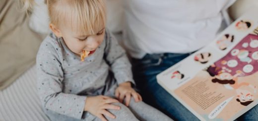 Cute toddler with ponytails enjoying a snack next to a book and parent. Cozy indoor scene.