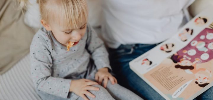 Cute toddler with ponytails enjoying a snack next to a book and parent. Cozy indoor scene.